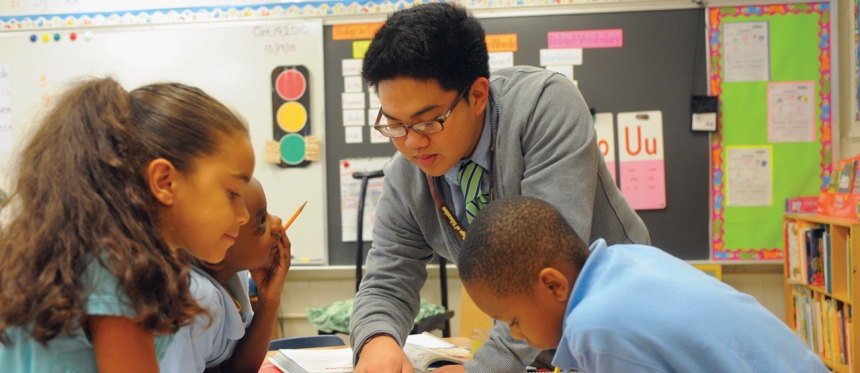 Elementary students sitting at desks are being assisted by a teacher.