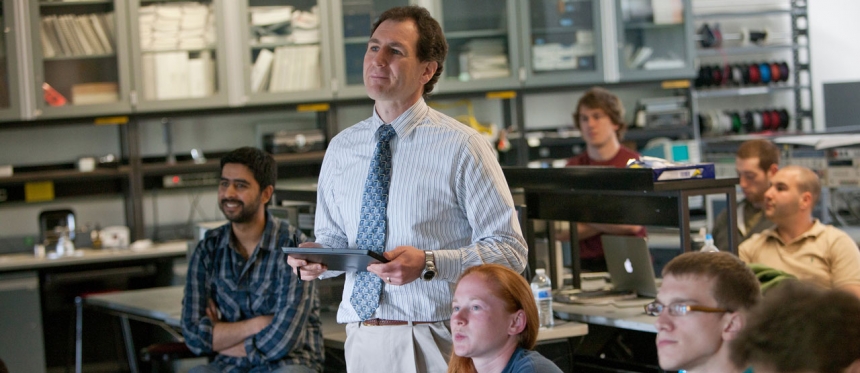 Professor with students in a classroom, appearing to be looking at a presentation off screen.