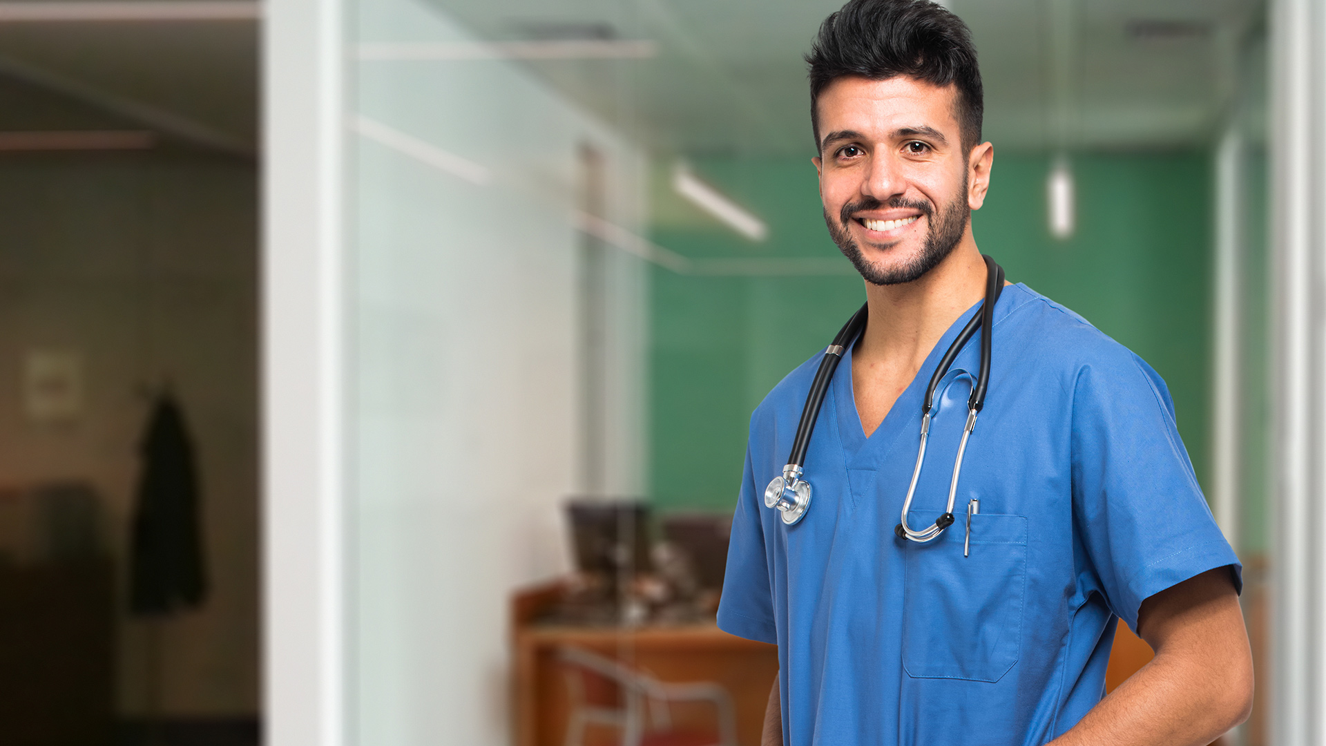 Smiling nurse with stethoscope, standing in hospital hallway.