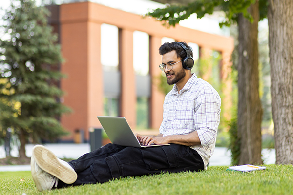 college student sitting under a tree with headphones on and an laptop open in front of him