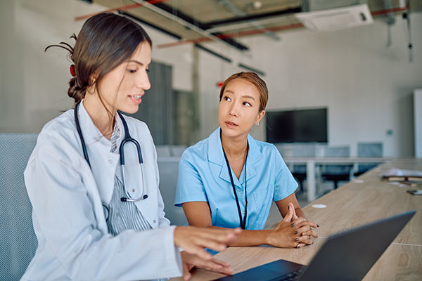 Nurse in lab coat on laptop sitting at a table and speaking with a student nurse.
