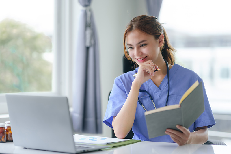 Nurse working on laptop, holding a book and pen.
