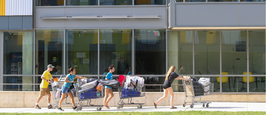 People push shopping carts filled with belongings outside a modern building. The scene conveys a sense of movement and purpose on a sunny day.