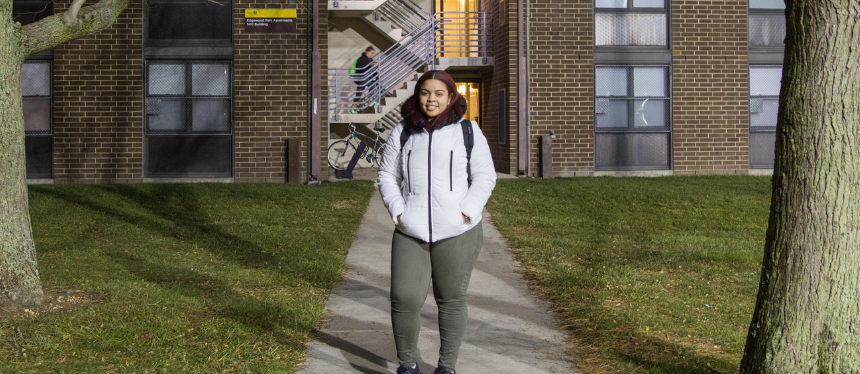 A person in a white jacket and backpack stands on a pathway at night, flanked by trees, in front of a brick building with a lit staircase.