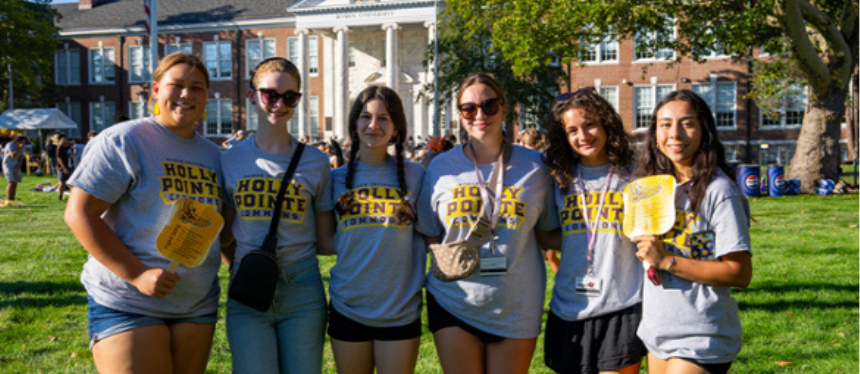 A group of six people stands smiling on a sunny campus lawn, wearing matching "Holly Pointe" shirts. A historic building with columns is in the background.