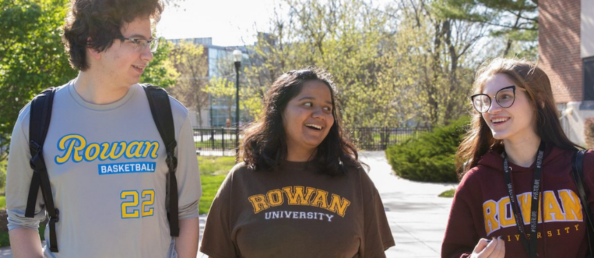 Three students walk and talk on a sunny campus path, wearing Rowan University apparel. They appear happy and engaged, with trees in the background.