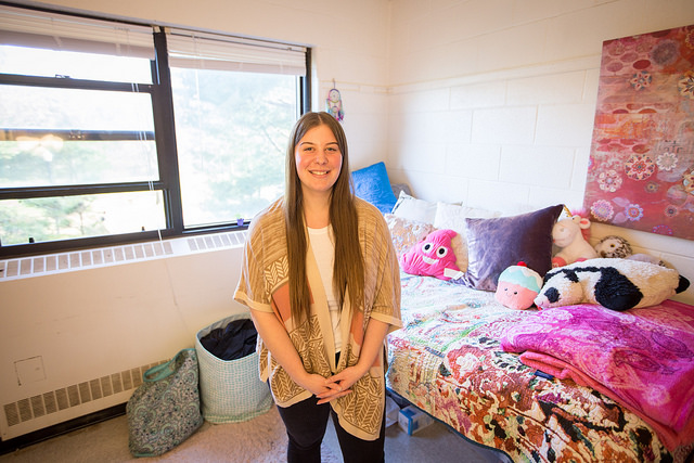 Young woman smiling in a cozy dorm room with colorful bedding, plush toys, and art on a white wall. Bright sunlight streams through the window.