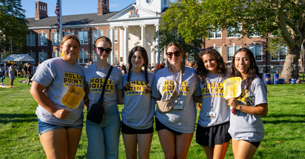 Six people stand smiling on a grassy campus lawn, wearing matching "Holly Pointe Commons" shirts. A historic brick building is visible in the background.