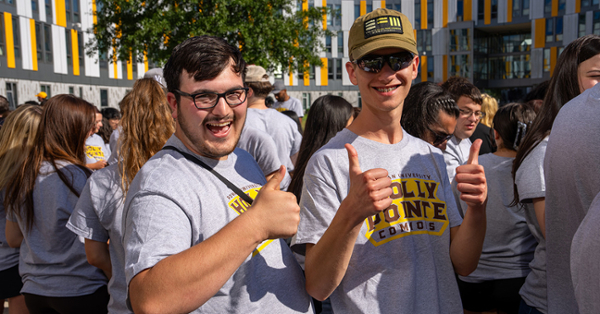 Two smiling individuals in matching gray "Welcome" shirts give thumbs up at an outdoor gathering. Surrounding crowd in similar attire, sunny day.