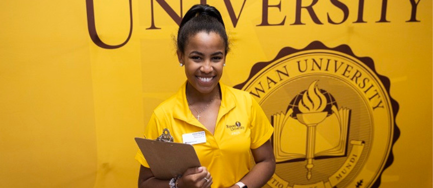 A woman in a yellow polo shirt smiles while holding a clipboard, standing in front of a large Rowan University banner, conveying a welcoming and cheerful atmosphere.