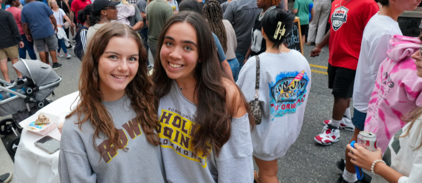 Two young women smile at the camera amid a crowded street festival. They wear casual sweatshirts, surrounded by people enjoying the lively atmosphere.