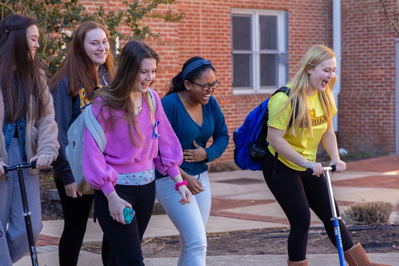 Five young women, smiling and laughing, walk and scooter together on a sunny day. They are outside near a brick building, displaying friendship and joy.