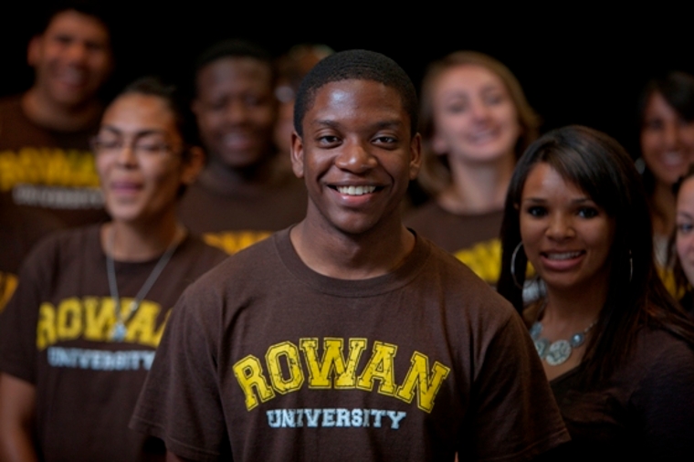 A group of smiling students wearing brown "Rowan University" T-shirts stand together. The central figure, a young man, is in focus, conveying a sense of camaraderie and school pride.