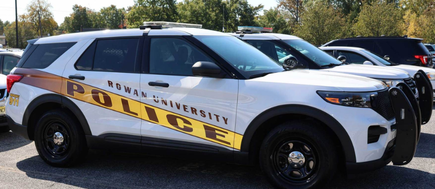 A white police SUV with "Rowan University Police" in bold letters is parked in a lot. Other vehicles are visible, and trees are in the background.