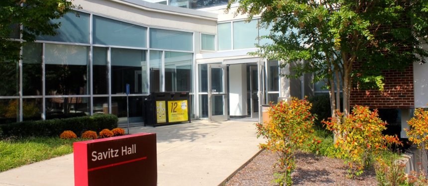 Entrance of Savitz Hall, featuring a red sign and modern glass facade. Surrounded by greenery and flowers on a sunny day, conveying a welcoming tone.
