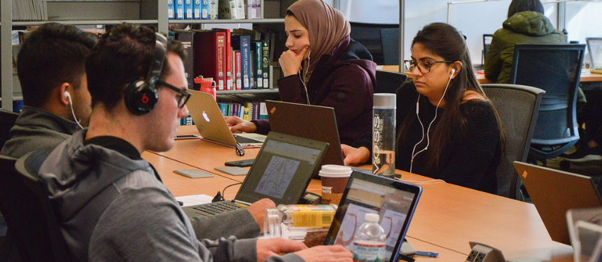 Students in a library study intently at laptops, surrounded by books. Two wear headphones, conveying focus. The atmosphere is quiet and studious.