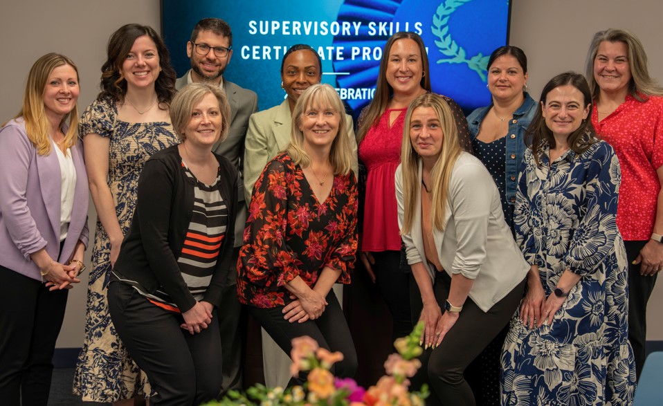 Supervisory Skills 2026 Cohort standing in front of a TV with "Supervisory Skills Certificate Program 2026 Celebration" displayed.