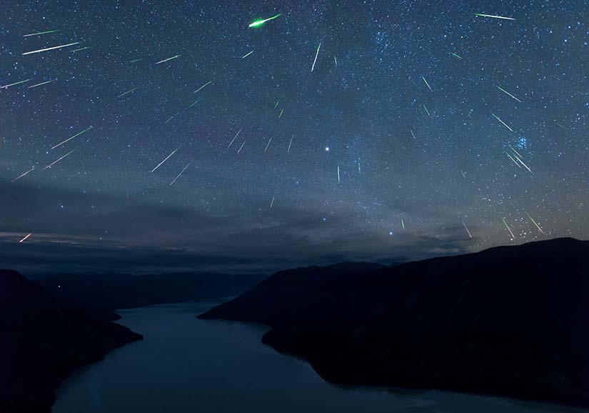 Meteors streak across a starry sky. On the horizon are mountains and a river.