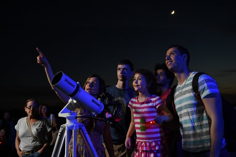 A group of people gathers around a telescope. A young girl in a pink and white striped shirt stands on a stool in front of the telescope. A woman is pointing to an object in the sky.