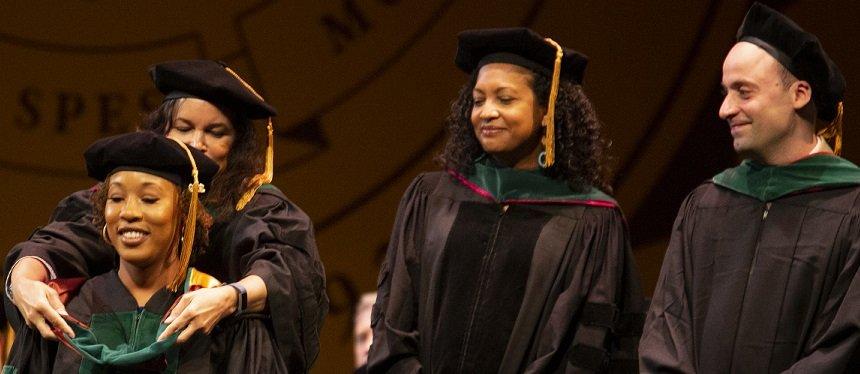 Four doctoral students are in their commencement regalia as one student is hooded on stage.