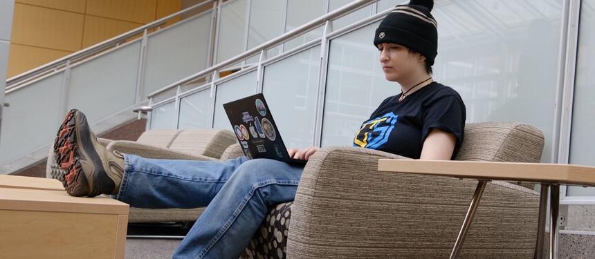 Person sitting casually with one leg raised, looking at a laptop covered with stickers.