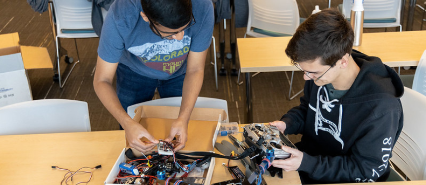 Two people work together at a table covered with wires, tools, and electronic parts. One person stands while holding a circuit board, and the other sits examining a mechanical component connected to several wires. The setting resembles a classroom or workshop with chairs and a box in the background.