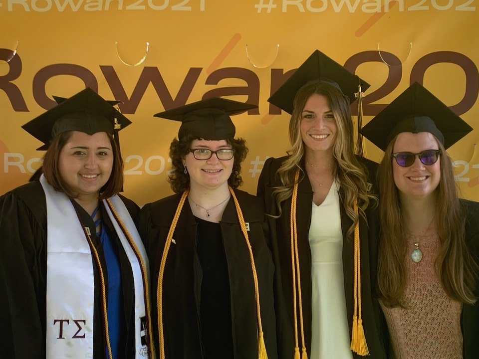 Four women standing in front of a Rowan banner wearing graduation attire.