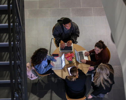 Overhead view of a group of students sitting around a round table with open laptops, collaborating in a campus lobby next to a staircase.
