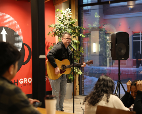 A musician playing an acoustic guitar and singing into a microphone near a speaker inside a coffee shop with audience members watching in the foreground.