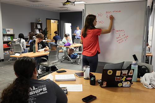 A tutor in a red shirt writes chemistry formulas on a whiteboard while students study at tables with laptops and books in a tutoring center.
