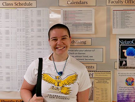 A smiling student, wearing a Flying First t-shirt, stands in front of a bulletin board. 