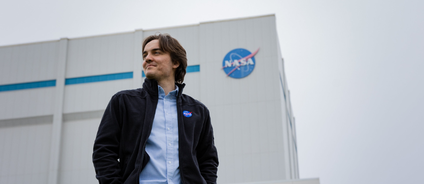 A person wearing a light blue shirt and a dark jacket with a NASA patch stands outside in front of a large white building featuring the NASA logo on its wall under an overcast sky.
