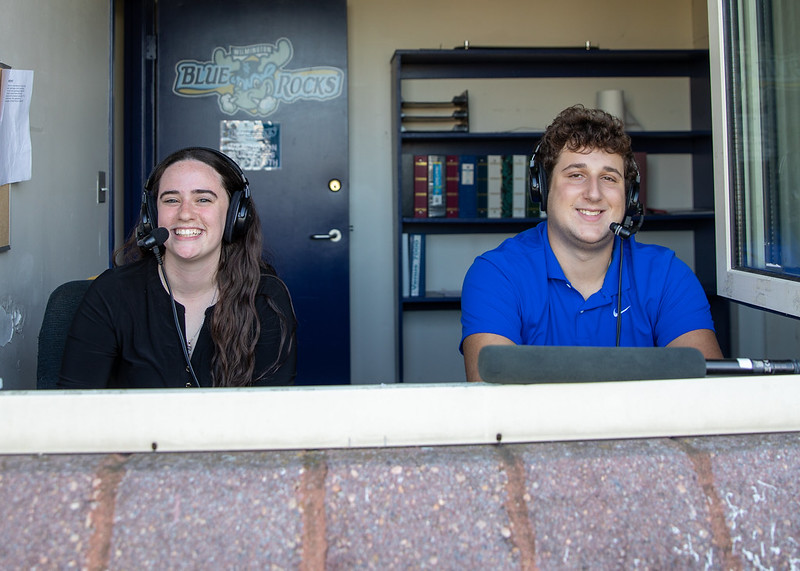 Two people sit at a desk with a laptop and microphone inside the Jaguar Radio Room, surrounded by shelves of books and binders.