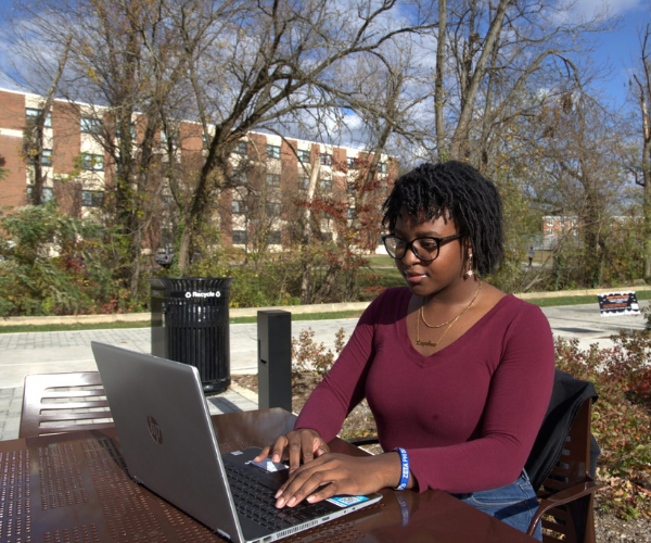 A student works on a laptop at an outdoor campus table on a sunny day.